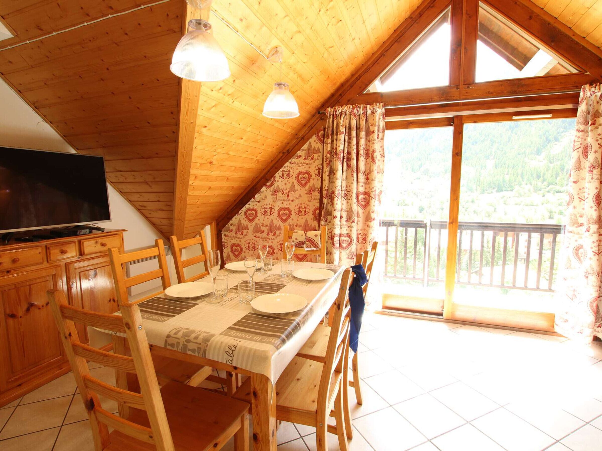 A cozy wooden dining area with a table set for four, a television on a wooden cabinet, and a balcony with a view of the forest.