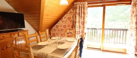 A cozy wooden dining area with a table set for four, a television on a wooden cabinet, and a balcony with a view of the forest.