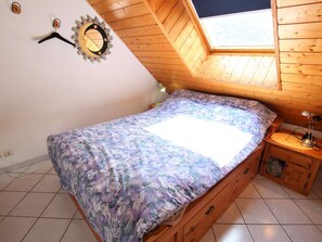 Une chambre avec un plafond en pente, un lit recouvert d’une couvre-lit à motifs floraux, une table de chevet avec une lampe et une horloge murale.