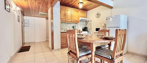 A dining area with a wooden table and chairs, a kitchen in the background, and a white refrigerator.