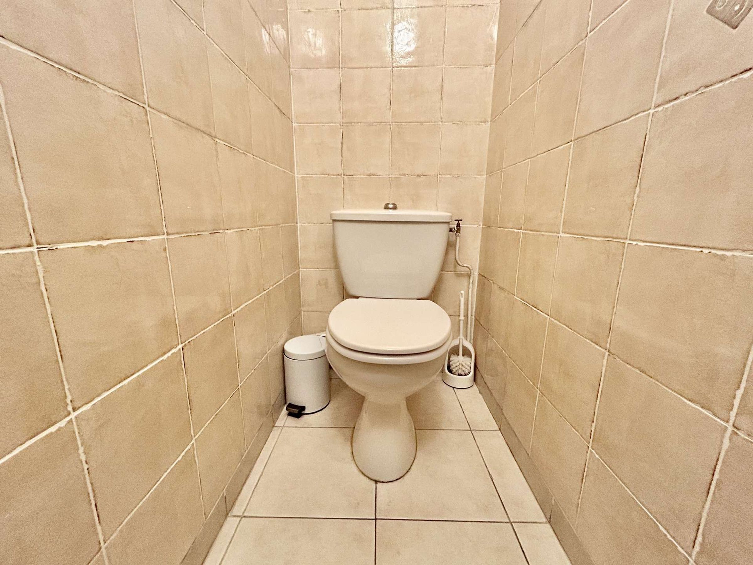 A small, tiled bathroom with a toilet, a trash bin, and a cleaning brush.