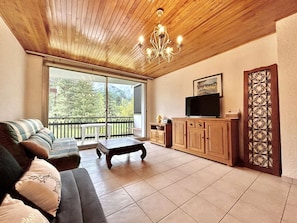 A living room with a sofa, a coffee table, a TV on a wooden cabinet, and a balcony with a view of trees.