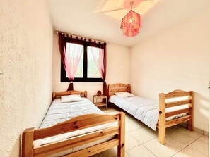 A room with two wooden beds, a window with curtains, a bedside table, and a hanging light fixture.
