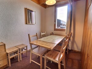 Ruokailutilassa on puinen pöytä ja tuolit, pieni ikkuna, jossa on verhot, sekä peili seinällä.