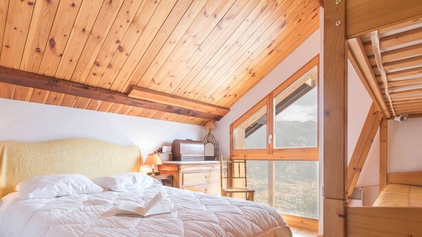 A bedroom with a wooden ceiling, a large bed with white bedding, a nightstand, a chair, and a window offering a mountain view.