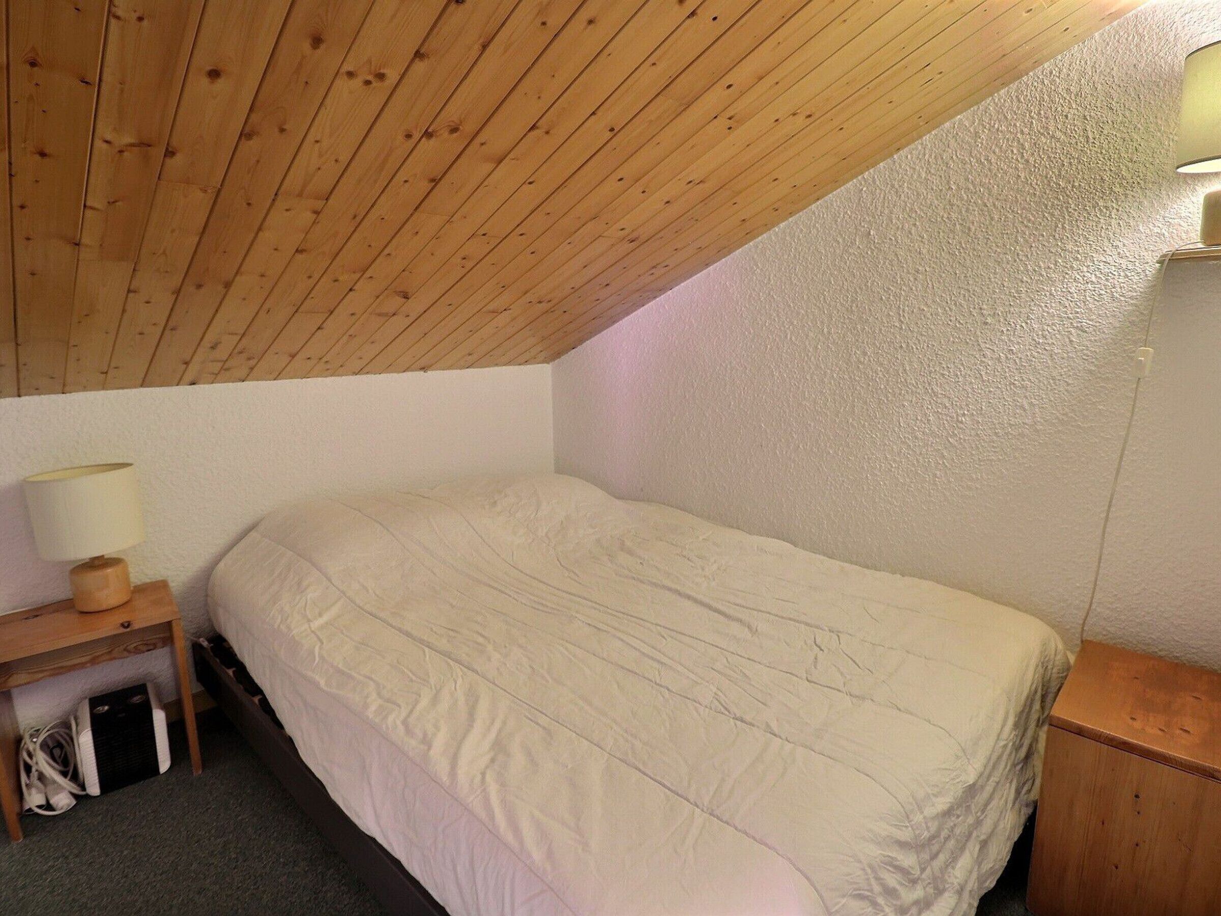 Ein kleines Zimmer mit einem Einzelbett, einer schrägen Decke, einem Nachttisch mit Lampe und einer Wandleuchte.