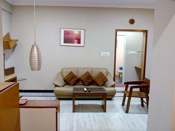 Una sala de estar con un sofá, una mesa de centro, un lámpara colgante, un cuadro en la pared y un espejo que refleja un baño.