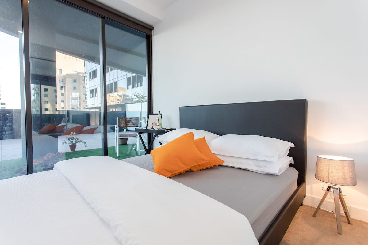 Une chambre d’hôtel avec un lit, une table de chevet, une lampe et une vue sur les bâtiments, visible à travers une porte vitrée.