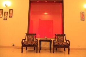 A room with two ornate chairs and a small table, a red-framed window, and wall-mounted lights.