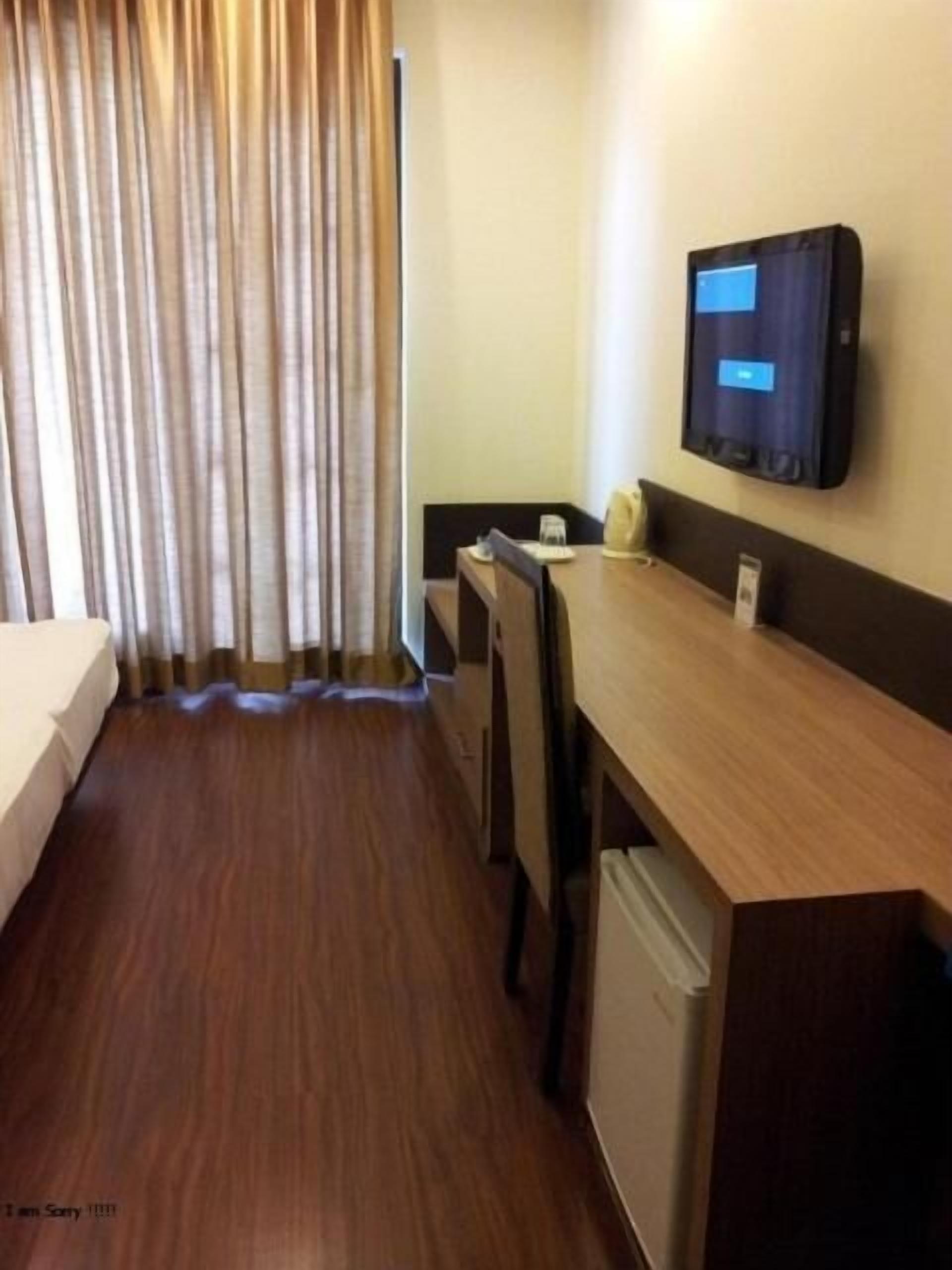 A hotel room with a bed, a desk, a chair, a small refrigerator, and a television mounted on the wall.