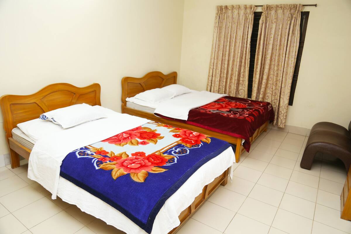 Two wooden beds with white and red floral bedspreads, a brown plastic stool, and a window with beige curtains.