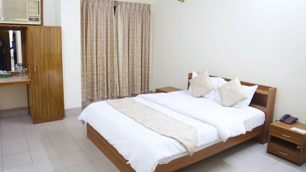 A hotel room with a wooden bed, white bedding, a bedside table with a phone, a small wooden cabinet, and a curtain. - Hotel Golden City (Sylhet)
