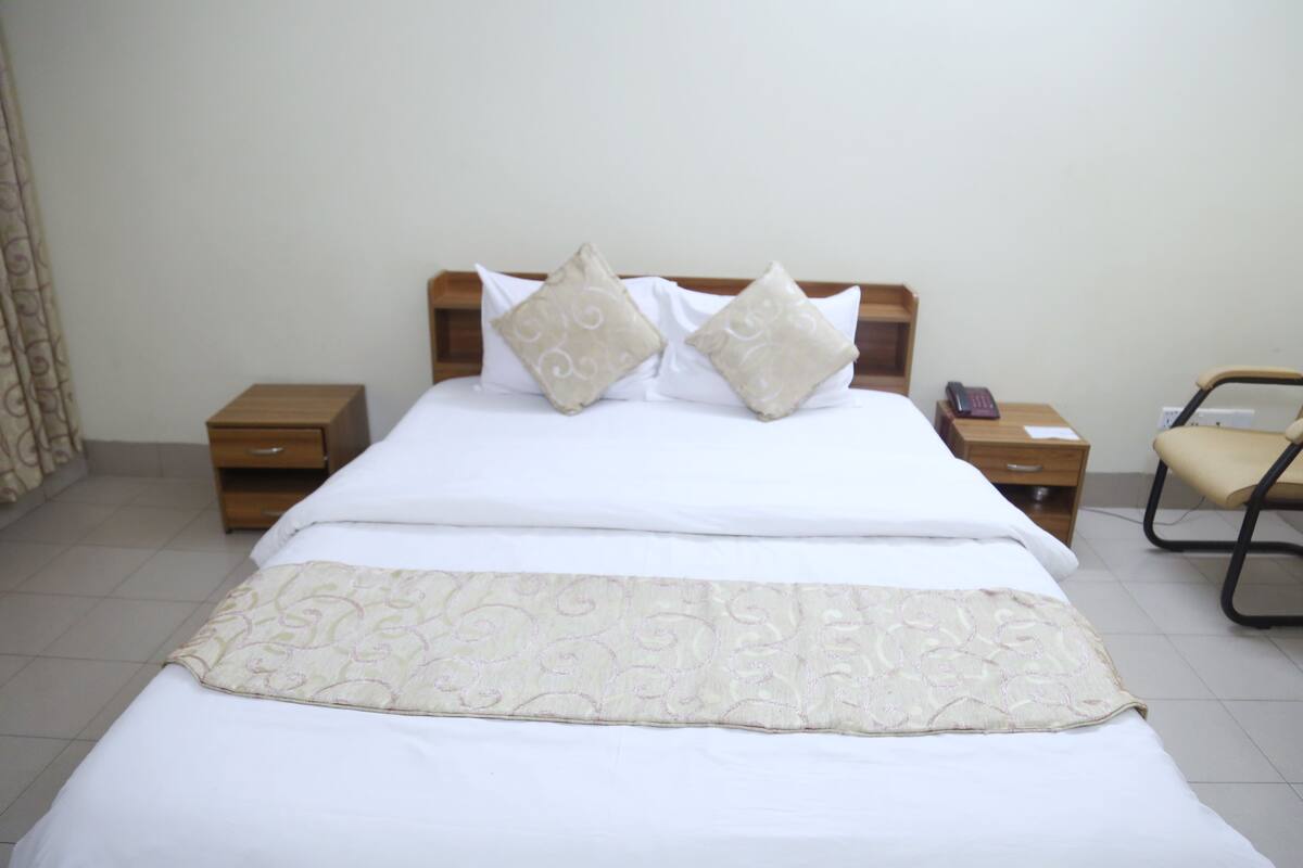A neatly made bed with a patterned bedspread, two pillows, and a bedside table with a phone and a lamp.
