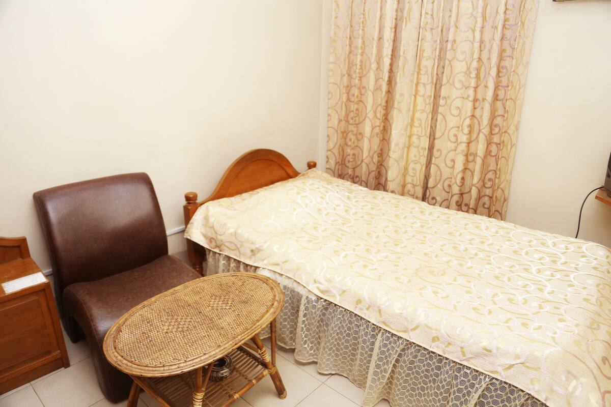 A small, neatly arranged bedroom with a single bed, a wooden chair, a small round table, and a curtain.