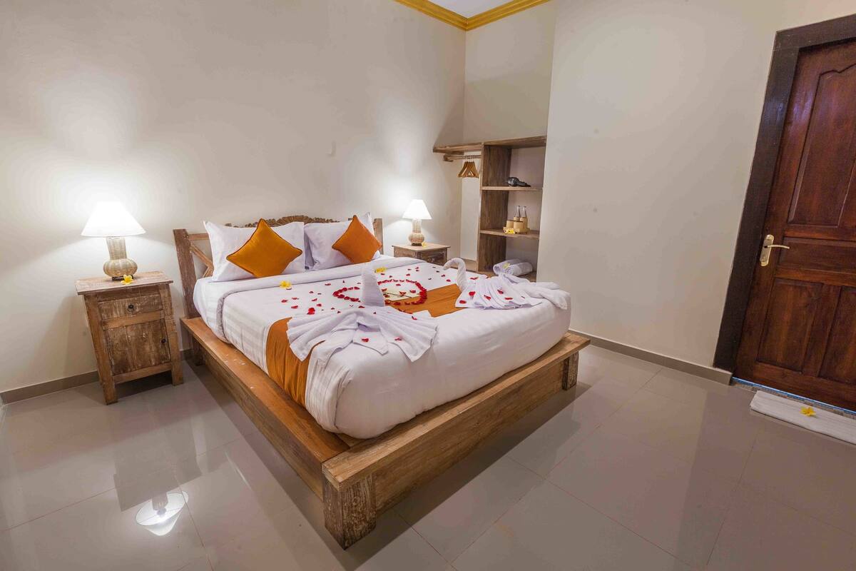 Un lit bien fait, avec du linge de lit blanc et des oreillers orange, deux tables de chevet avec des lampes, une tête de lit en bois et une porte en bois.