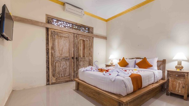 A bedroom with a wooden bed, white bedding, and orange pillows. There is a wooden nightstand with a lamp and a small table with a decorative item. - Vimala Ubud Hotel (Ubud)