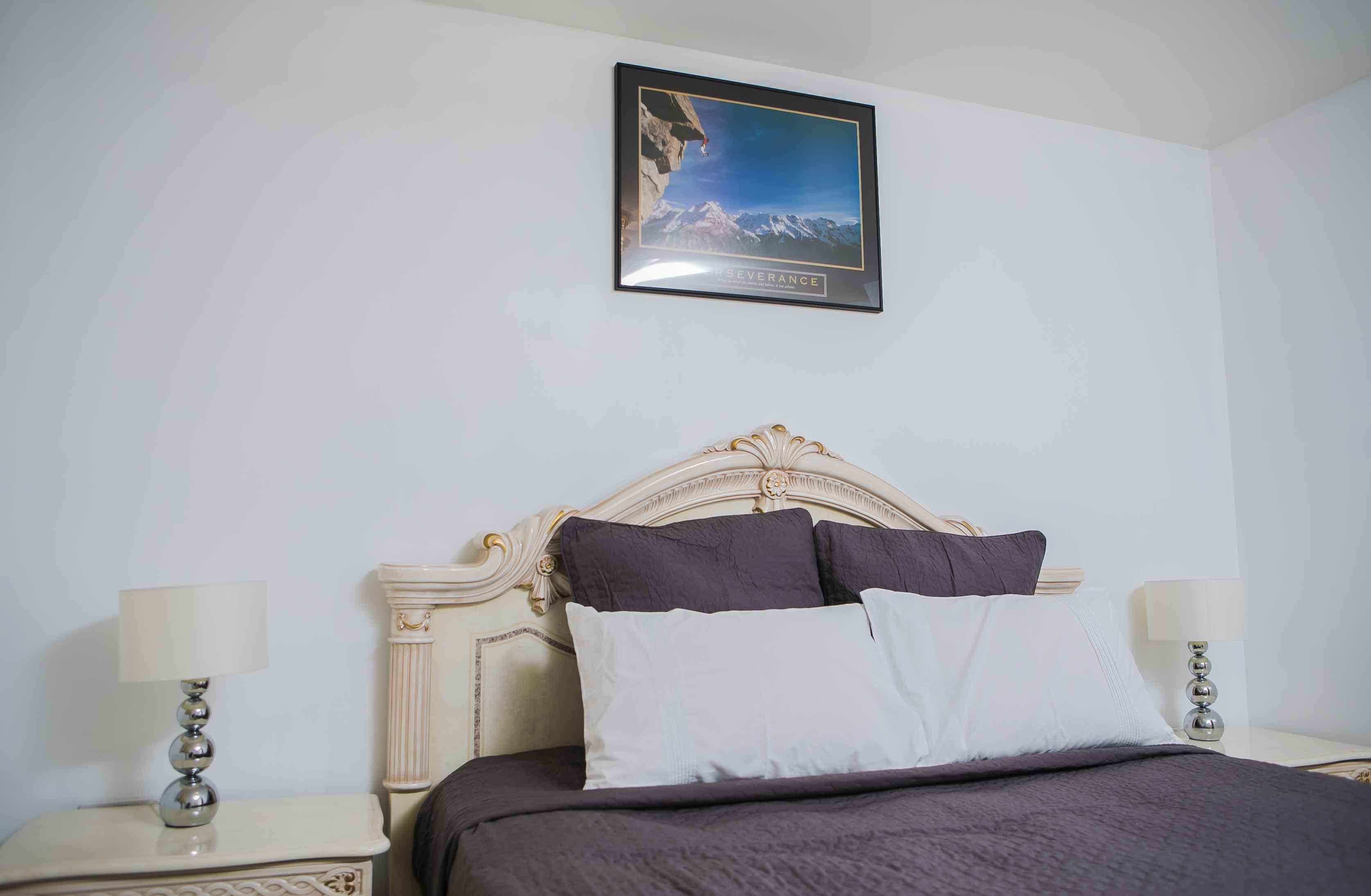A neatly made bed with a white and gray color scheme, a framed picture of a mountain on the wall, and two bedside lamps.