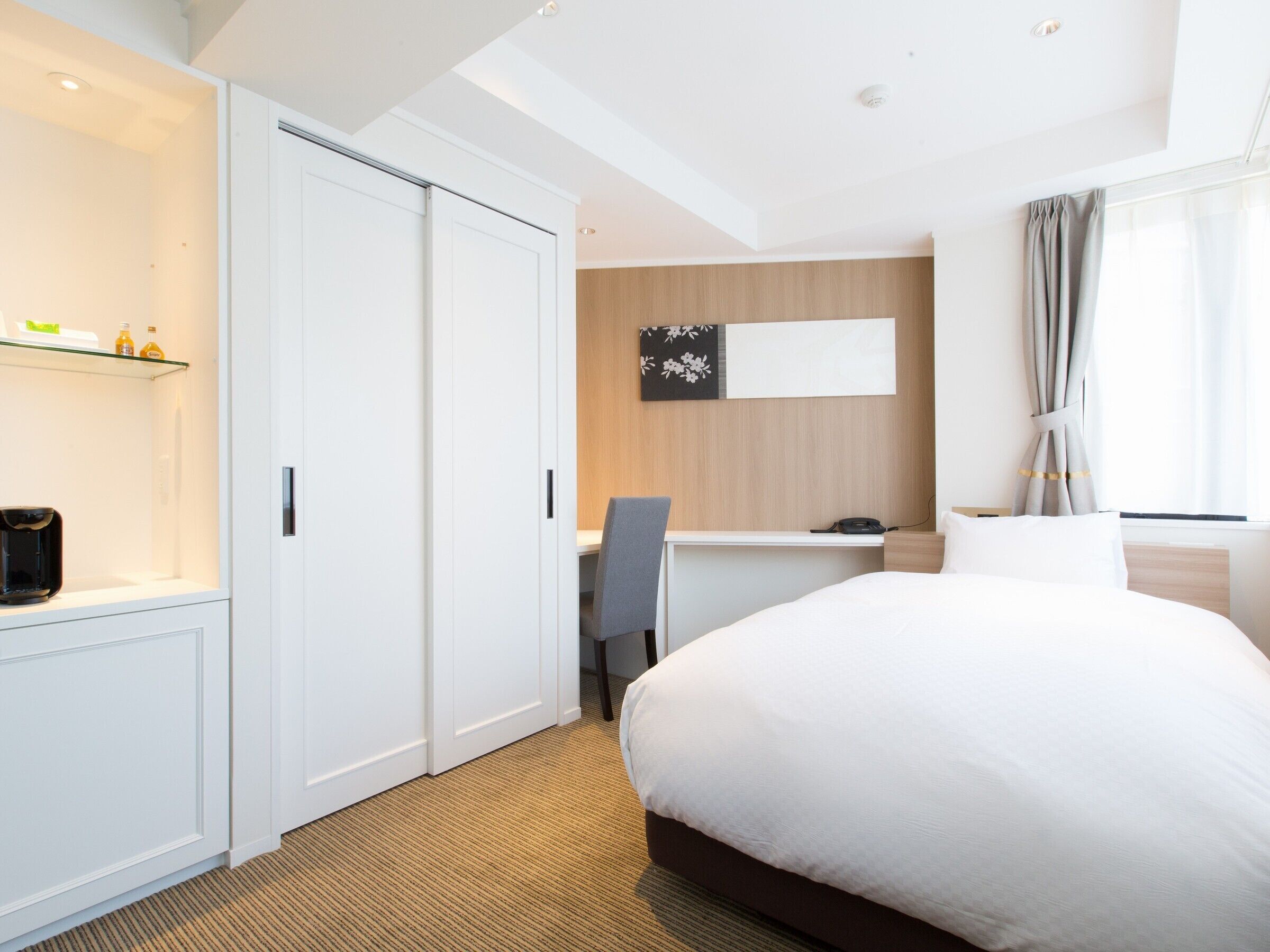 A hotel room with a bed, a desk, a chair, a wardrobe, and a shelf with items.