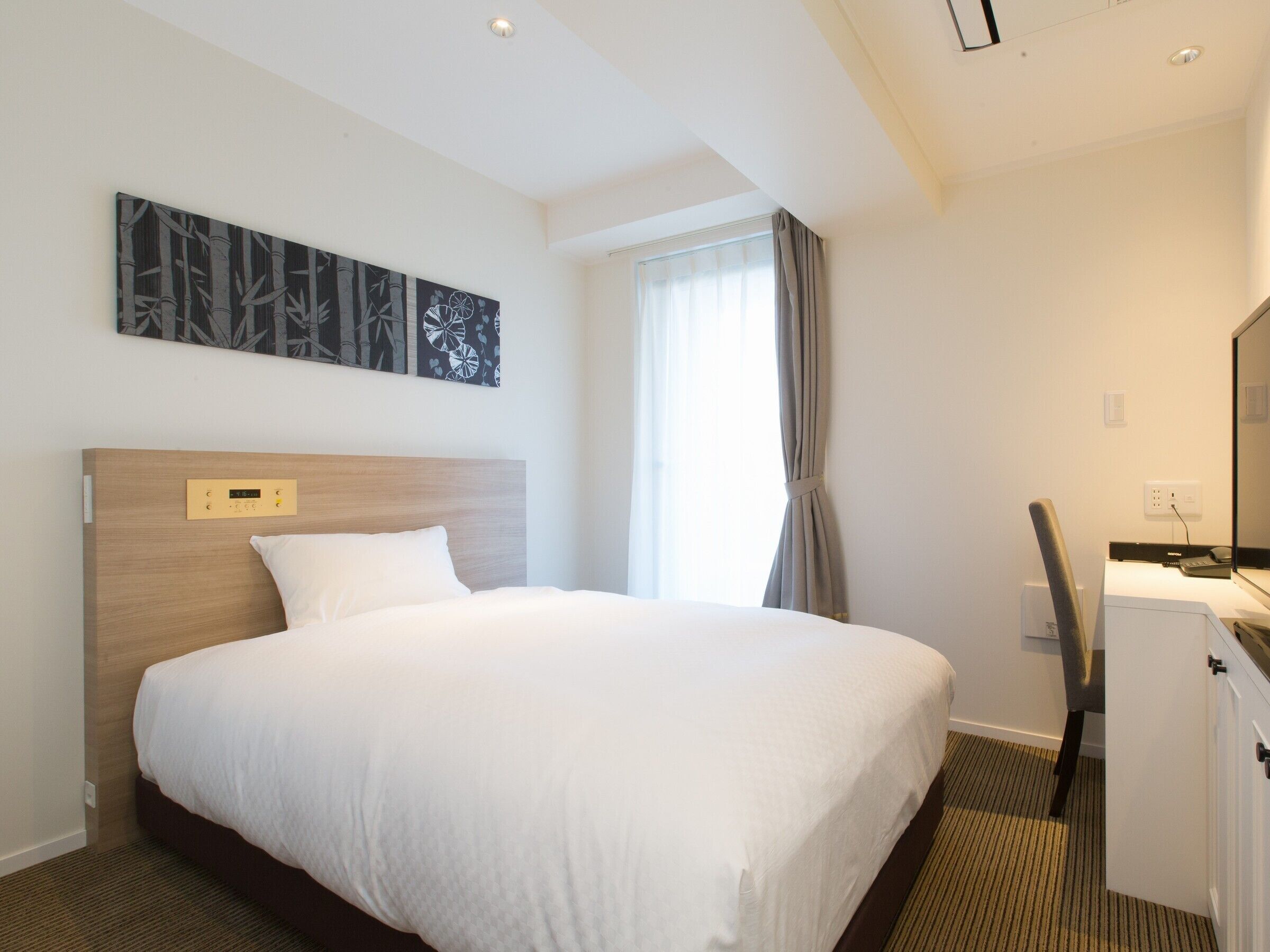 A hotel room with a bed, a desk, a chair, a window with curtains, and a wall-mounted artwork.