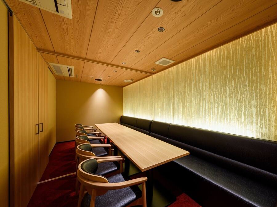 A conference room with a long table and several chairs, a bench along the wall, and a wooden ceiling with recessed lighting.