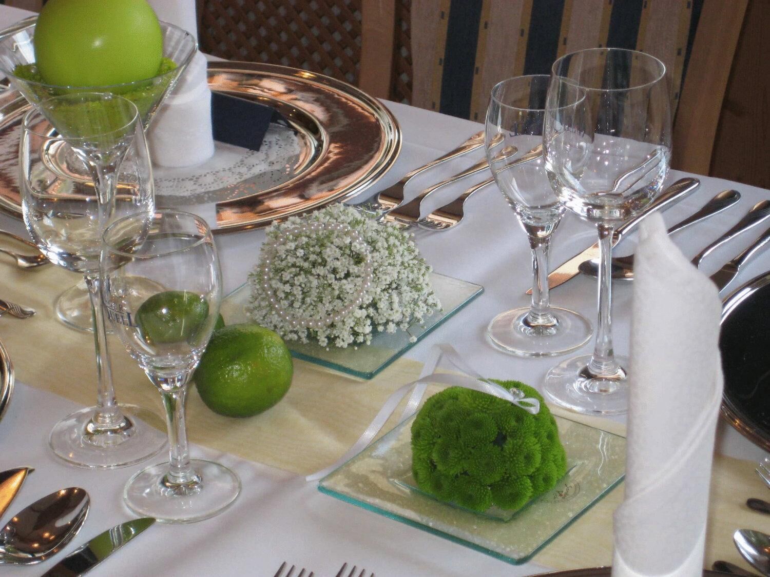 Una mesa dispuesta con cristalería transparente, cubiertos y objetos decorativos, incluyendo una fruta verde, un pequeño ramo y un objeto esférico verde.