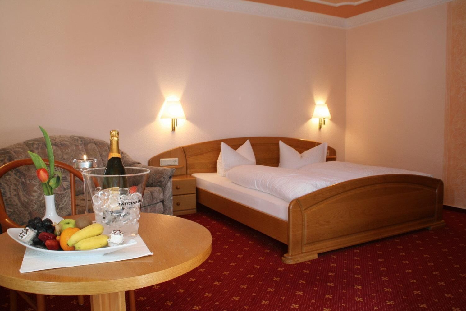 A hotel room with a wooden bed, two bedside lamps, a round table with fruit and a bottle of champagne, and a floral-patterned carpet.