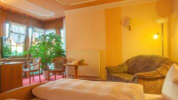 Habitación de hotel con cama, sofá, mesa de comedor con sillas, una planta en maceta y ventana con cortinas.