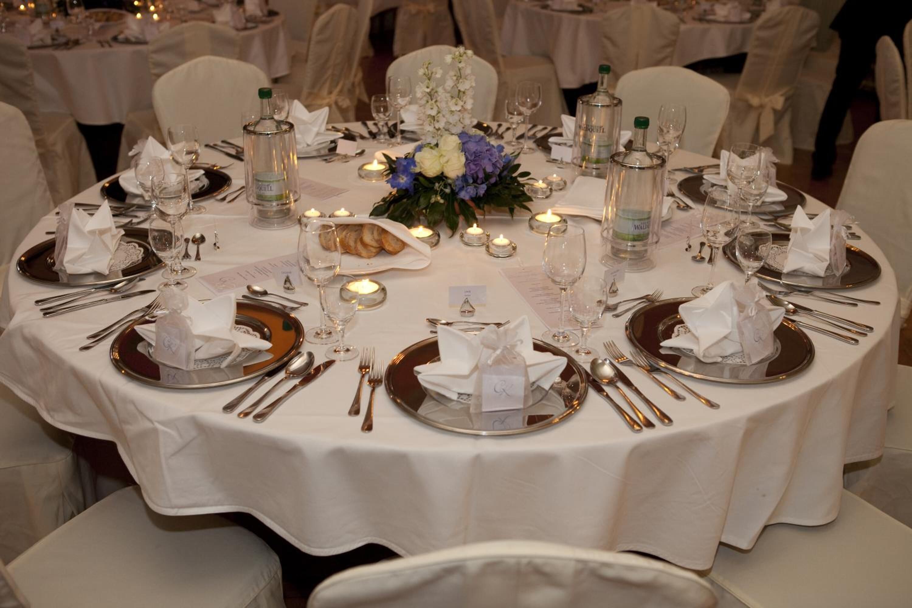 Un tavolo rotondo apparecchiato per un evento formale, con tovaglioli bianchi, sottopiatti dorati e posateria d'argento.