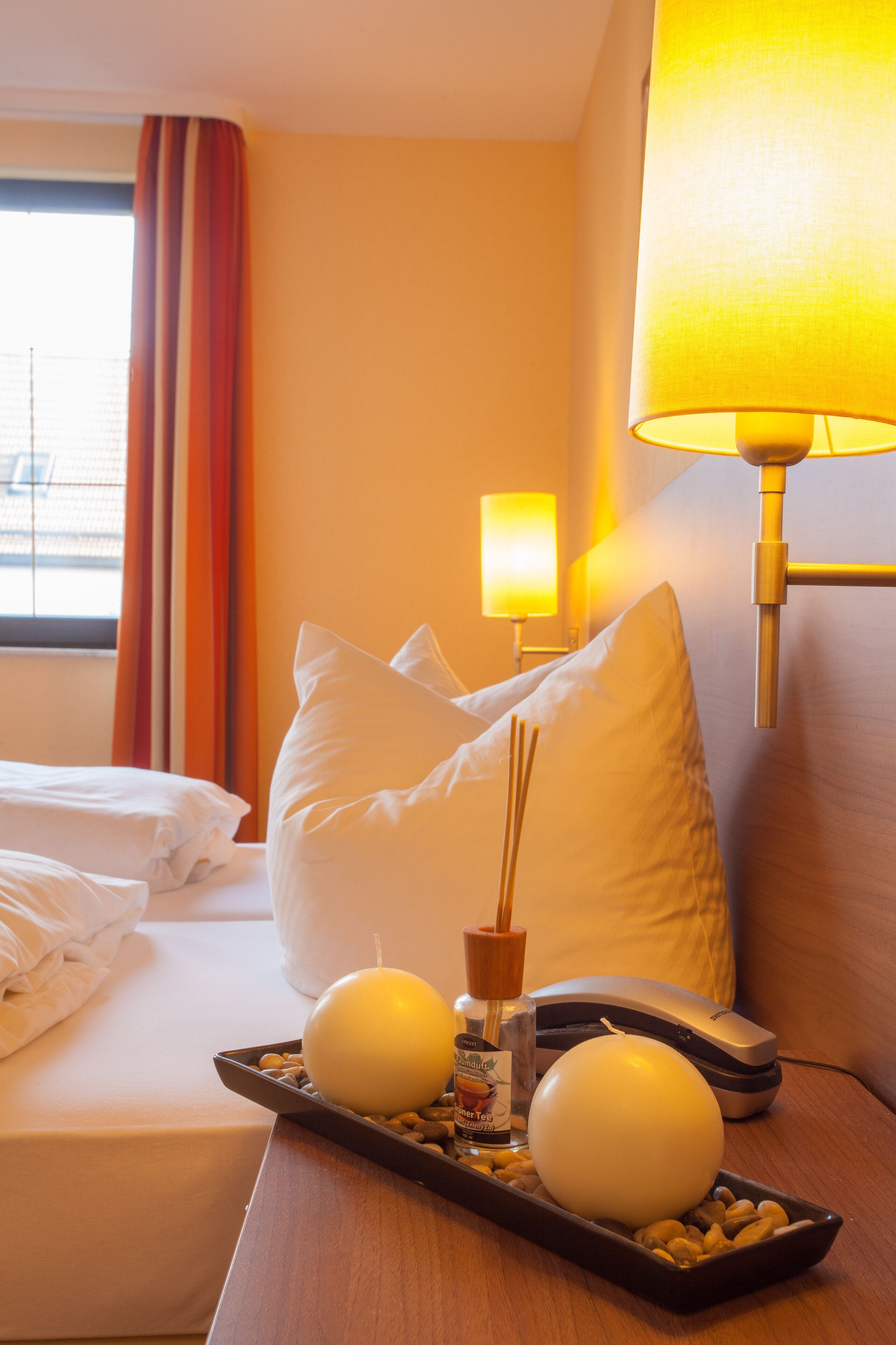 A hotel room with a neatly made bed, a bedside lamp, a remote control, and a tray with candles and a bottle.