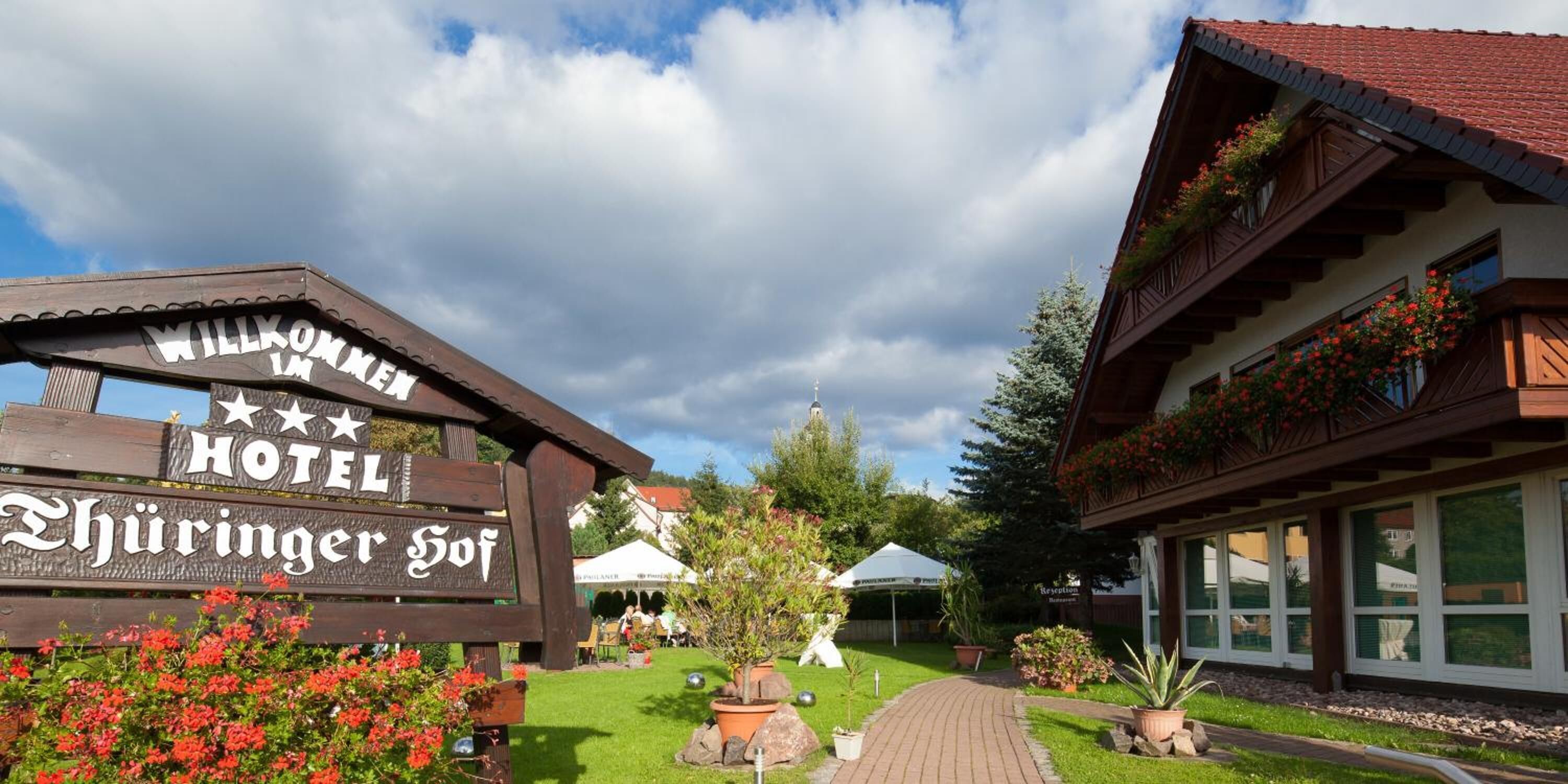 A wooden sign with "Willkommen Hotel" and "Hüringer Hof" text, a pathway leading to a building with large windows and flower boxes, and a clear sky.
