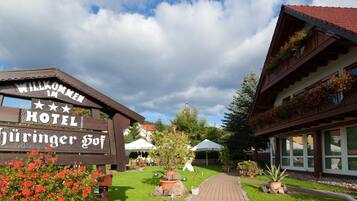 A wooden sign with "Willkommen Hotel" and "Hüringer Hof" text, a pathway leading to a building with large windows and flower boxes, and a clear sky.