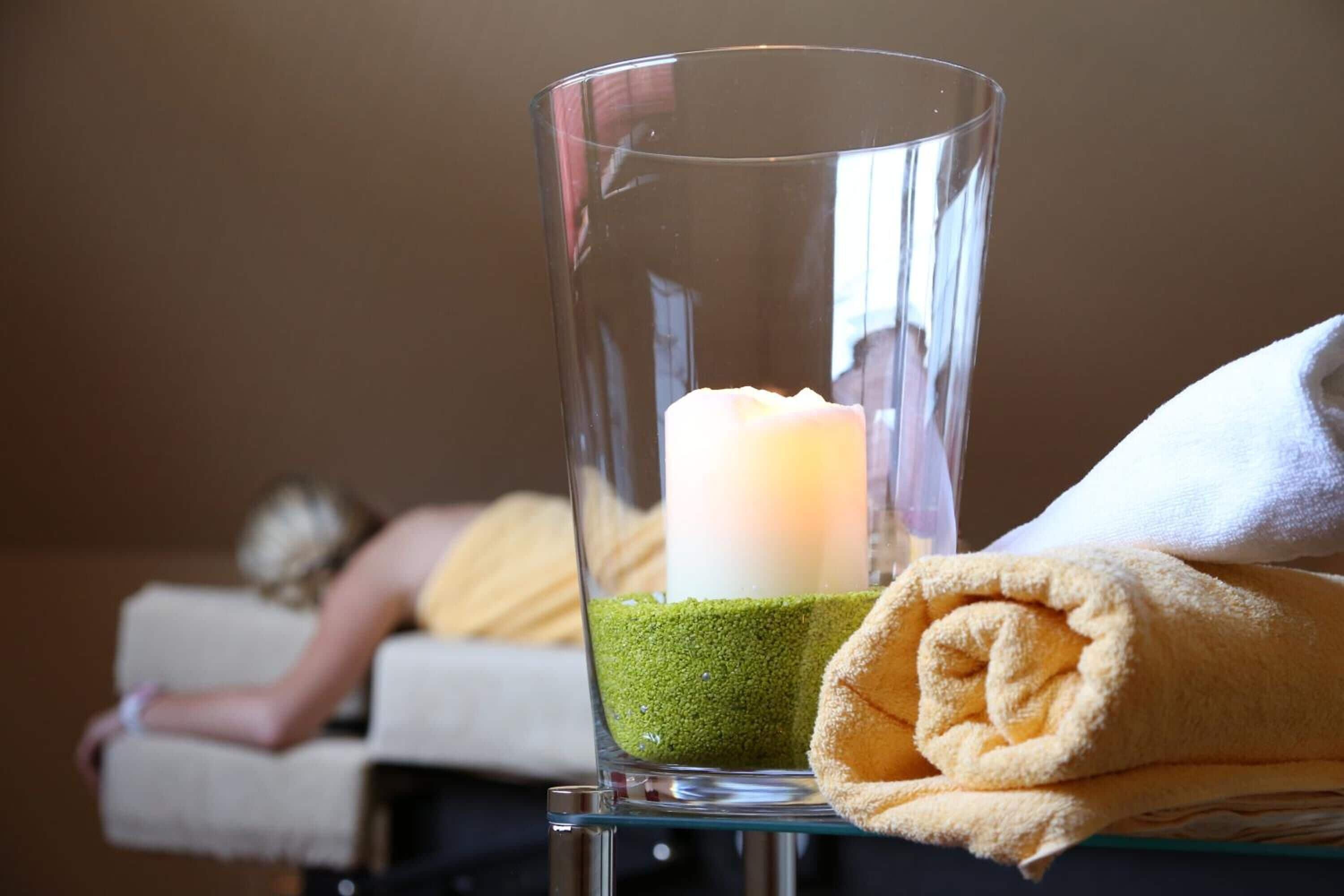 A glass candle holder with a candle and a rolled towel on a table.