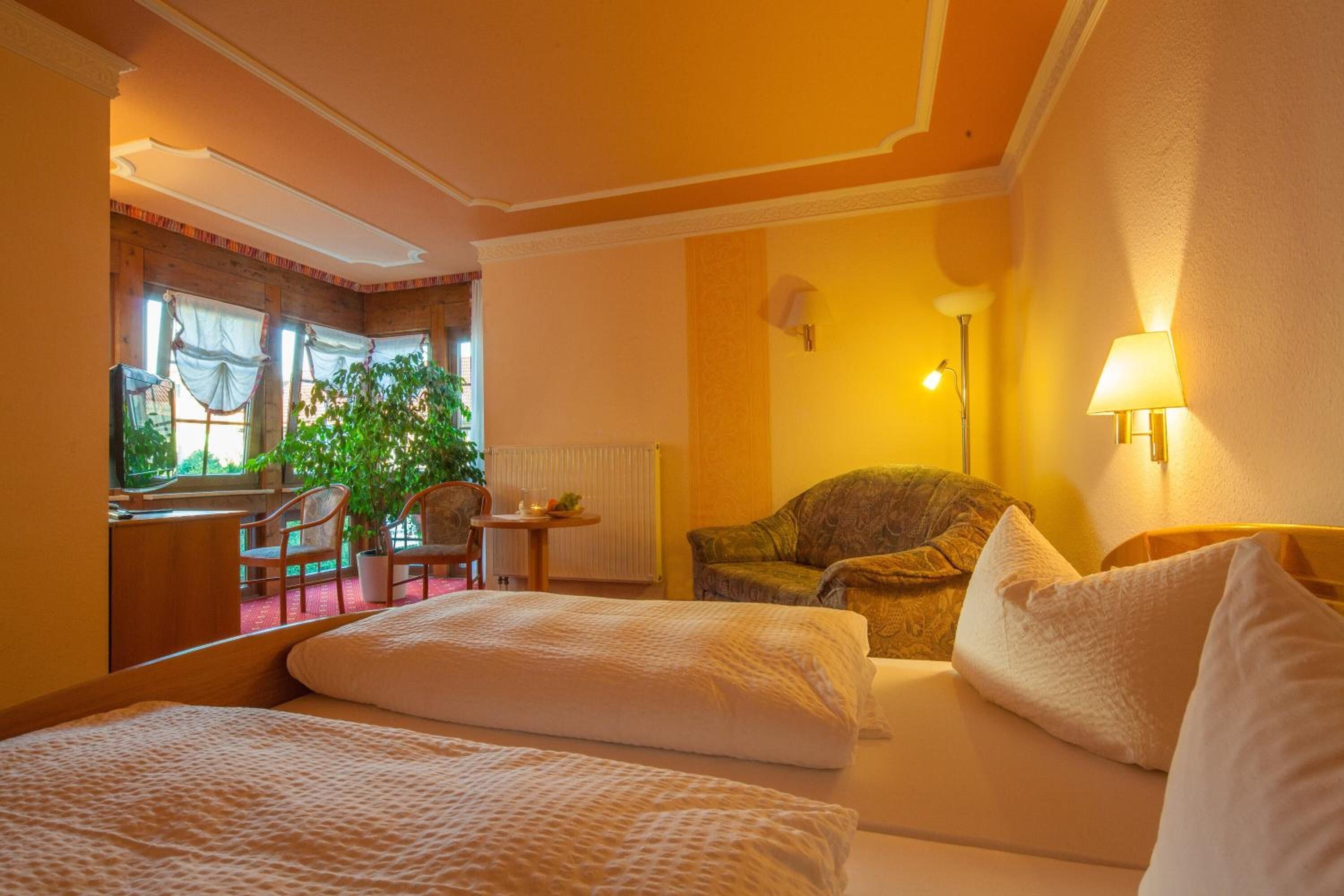 A hotel room with two beds, a sitting area with a chair and a table, a potted plant, and a view of the outdoors through a window.
