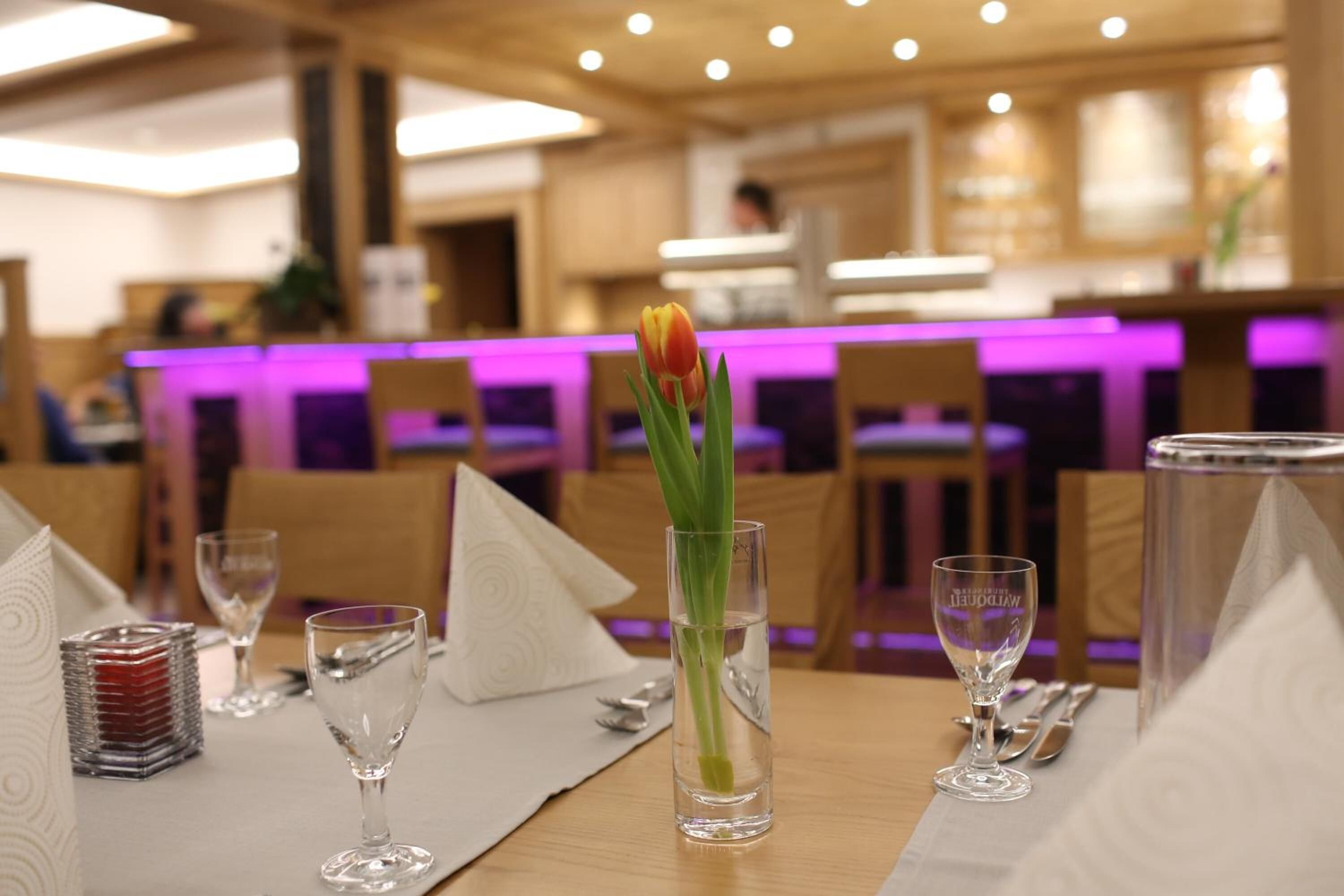 Une table à manger dressée avec un tulipe dans un vase en verre, des verres à vin et des couverts.