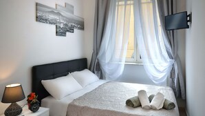 Ein ordentlich bezogenes Bett mit weißer Decke und Kissen, ein Nachttisch mit Lampe und Uhr sowie ein an der Wand befestigtes Fernsehgerät.