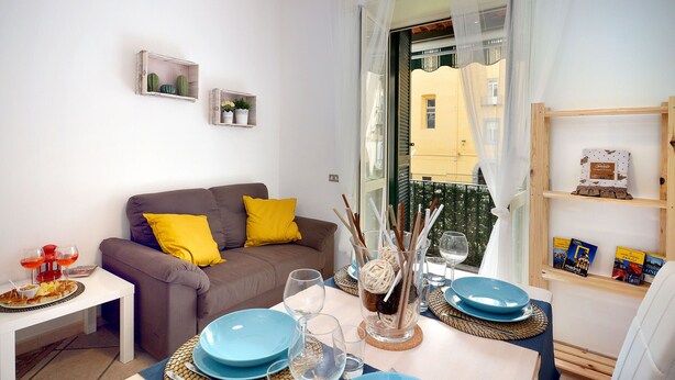 Ein Wohnzimmer mit einer Couch, einem Esstisch gedeckt mit blauen Tellern und Gläsern, sowie einem Bücherregal mit Büchern und Dekorationsobjekten.