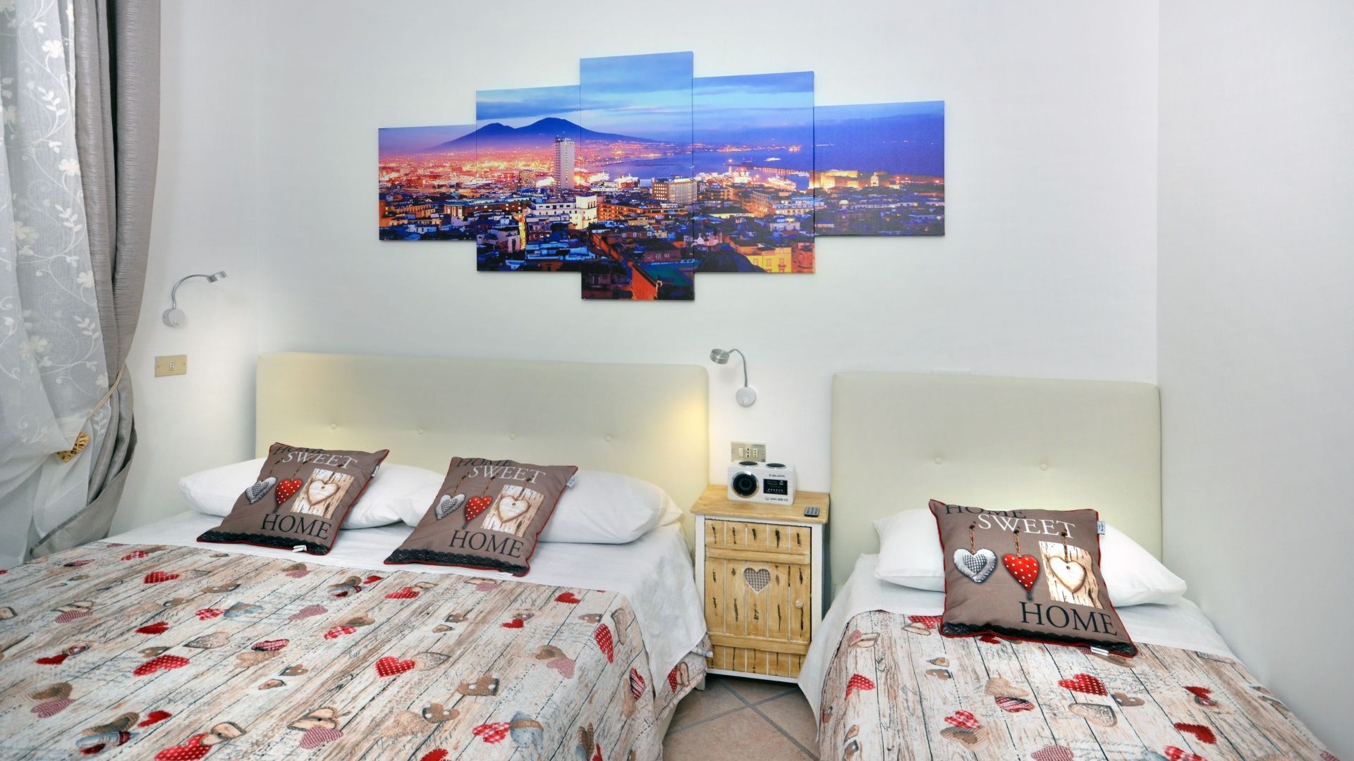 Ein Hotelzimmer mit zwei Betten, jeweils mit einem Bettbezug und Kissen mit Herzmuster. Zwischen den Betten befindet sich ein Nachttisch mit Radio und Lampe. Darüber hängt ein großes Stadtbild.