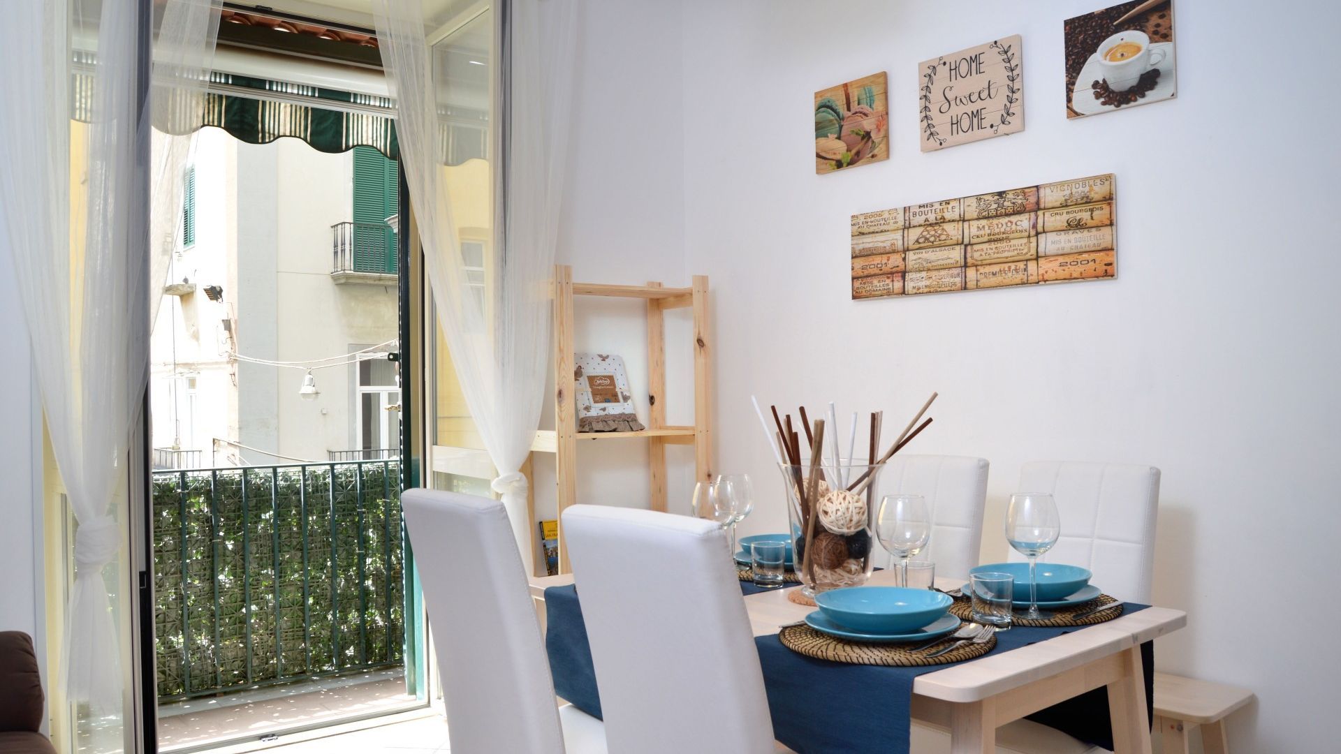 A dining area with a table set for two, a wooden shelf with decorative items, and a view of the outside balcony.