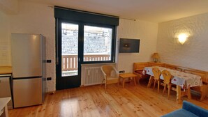Un espacio habitable compacto con kitchenette, una zona de comedor con mesa y sillas, y un ventanal con vista a un edificio de ladrillo.