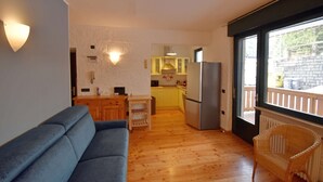 Una sala de estar con un sofá, una silla de mimbre, una zona de cocina con refrigerador y vistas a través de un ventanal.