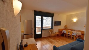 Una habitación acogedora con piso de madera, una mesa de comedor con sillas, un sofá, un refrigerador y un ventanal corredizo que da a un balcón.