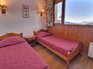 A small, cozy room with two wooden beds, a nightstand, a wall-mounted lamp, and a window with a view of mountains. - Charming 2-Room Apartment with Wi-Fi - Méribel (Méribel)