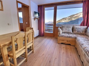 A wooden interior with a dining table, chairs, a sofa with patterned cushions, a television on a wall, and a large window with a mountain view. - Charming 2-Room Apartment with Wi-Fi - Méribel (Méribel)