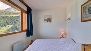 A bedroom with a bed, a window with a view of a forest and mountains, a wall-mounted lamp, and a framed picture. - Cozy Apartment in Méribel, 4 pers, Pets allowed (Méribel)