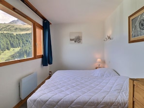 A bedroom with a bed, a window with a view of a forest and mountains, a wall-mounted lamp, and a framed picture. - Cozy Apartment in Méribel, 4 pers, Pets allowed (Méribel)
