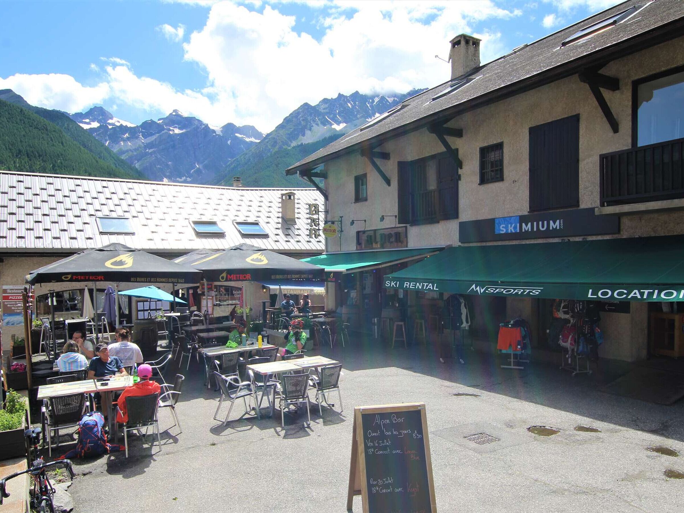 An outdoor café with tables and chairs, a chalkboard menu, and a mountain backdrop.