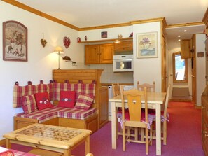 A cozy living room with a sofa, a wooden table, and a chessboard. There is a kitchen area with wooden cabinets and a microwave.