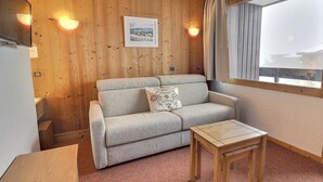 A cozy room with a wooden wall, a sofa, a small table, and a window with curtains. - Ski-in studio, pets allowed, sleeps 4 (Méribel)