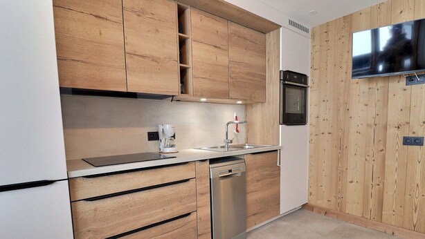 A modern kitchen with wooden cabinets, a built-in oven, a dishwasher, and a flat-screen TV mounted on the wall.