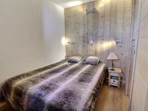 A bedroom with a wooden wall, a bed with a blanket, a bedside table with a lamp, and a small drawer unit.